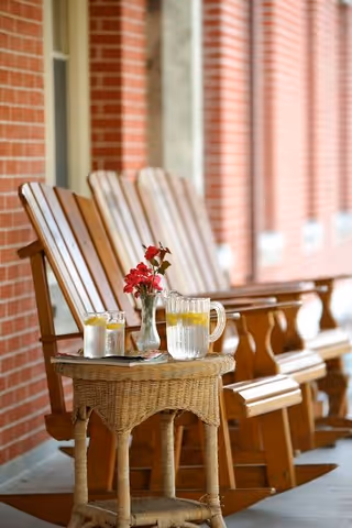 A row of wooden rocking chairs on a porch with a small wicker table in front holding a pitcher of water with lemon slices, two glasses of water, and a small vase with red flowers. The background shows a brick wall with windows.
