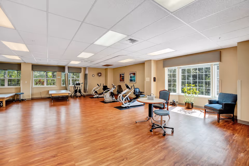 Bright, spacious interior activity/fitness room with hardwood floors, recumbent exercise bikes, seating, and large windows.
