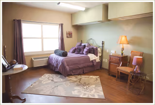 A cozy bedroom with a neatly made bed featuring purple bedding and multiple pillows. The room has a large window with curtains, a wooden nightstand with a lamp, a chair, a small round table with a lamp, and a floral patterned area rug on wooden flooring.