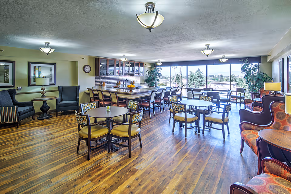 A spacious senior living common area with wooden flooring, multiple round tables with chairs, a long counter with bar stools, large windows letting in natural light, and comfortable armchairs along the walls.