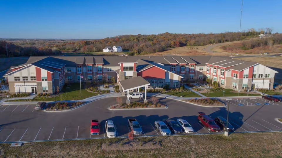 Aerial view of a large, modern senior living facility building with a covered entrance and multiple windows. Several cars are parked in the parking lot in front of the building. The facility is surrounded by open land and trees with autumn foliage under a clear blue sky.