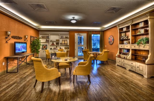 A cozy common area with wooden flooring and warm orange walls. The room features several yellow cushioned chairs arranged around circular tables. There are built-in white shelves with books and decorative items on the far wall, and a large wooden cabinet with more books and plants on the right. A flat-screen TV is mounted on the left wall above a wooden console table. The room is well-lit with ceiling lights and natural light coming through a glass door and window at the back.