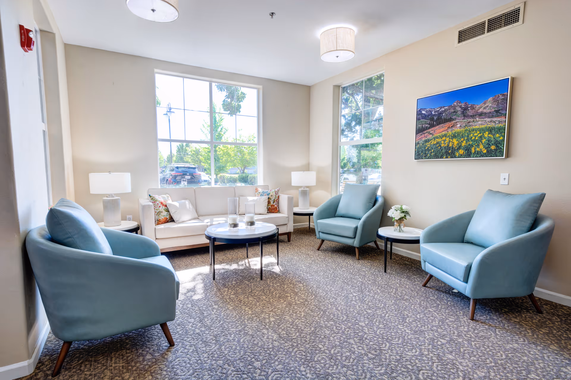 A bright and cozy living room with large windows letting in natural light. The room features a white sofa with decorative pillows, three light blue armchairs, two side tables with lamps, a round coffee table with candles, and a wall-mounted TV displaying a scenic mountain and flower landscape.