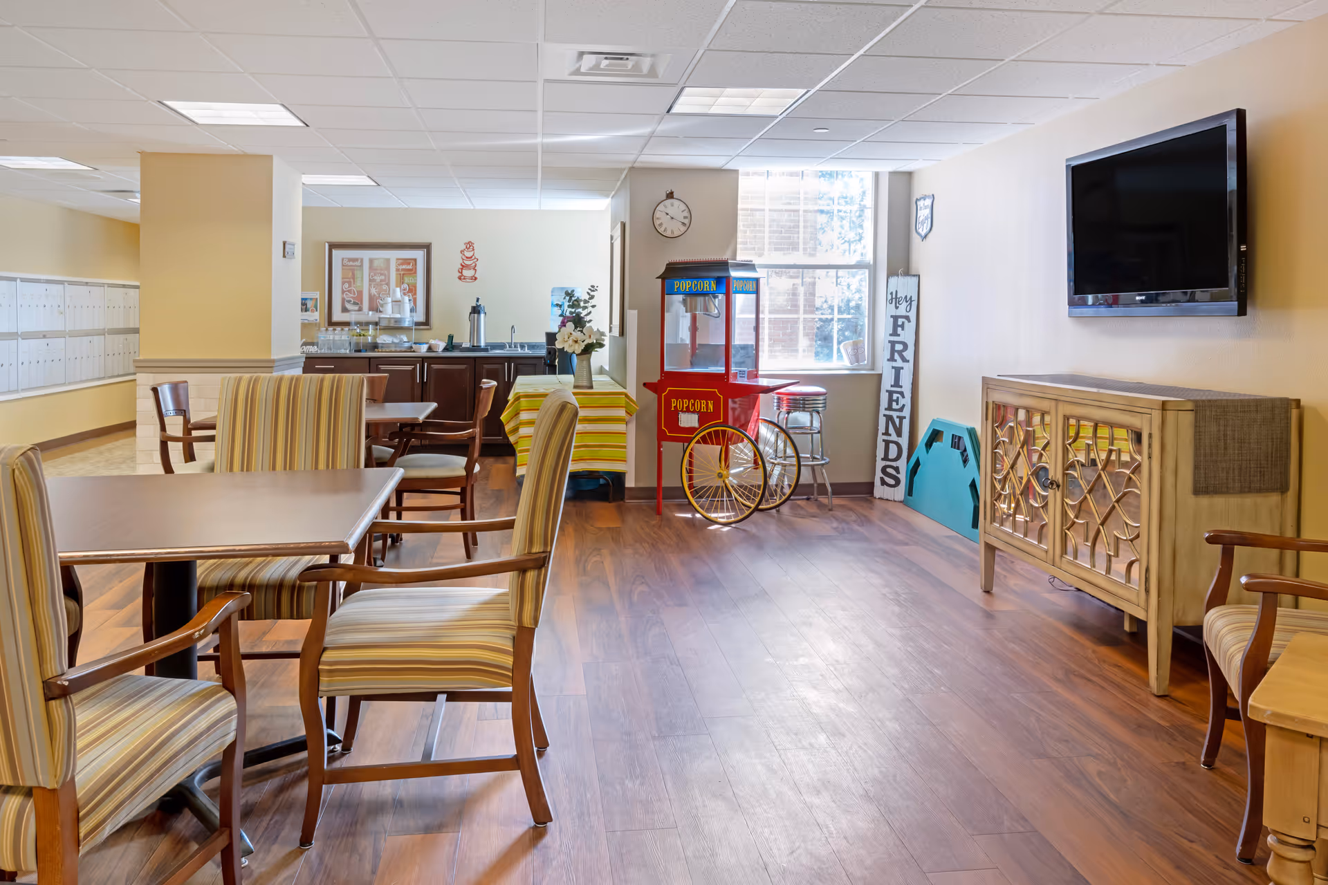 A cozy common area in a senior living facility with wooden flooring, several tables and striped cushioned chairs, a popcorn machine near a window, a wall-mounted TV, and a cabinet. There is a small kitchenette area with a coffee station and a table covered with a yellow striped cloth. A decorative sign reads 'Hey Friends'.