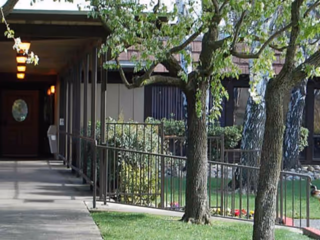Outdoor view of a walkway leading to the entrance of a building with a wooden door and circular window. The walkway is bordered by a metal railing and surrounded by green grass, trees, and shrubs.