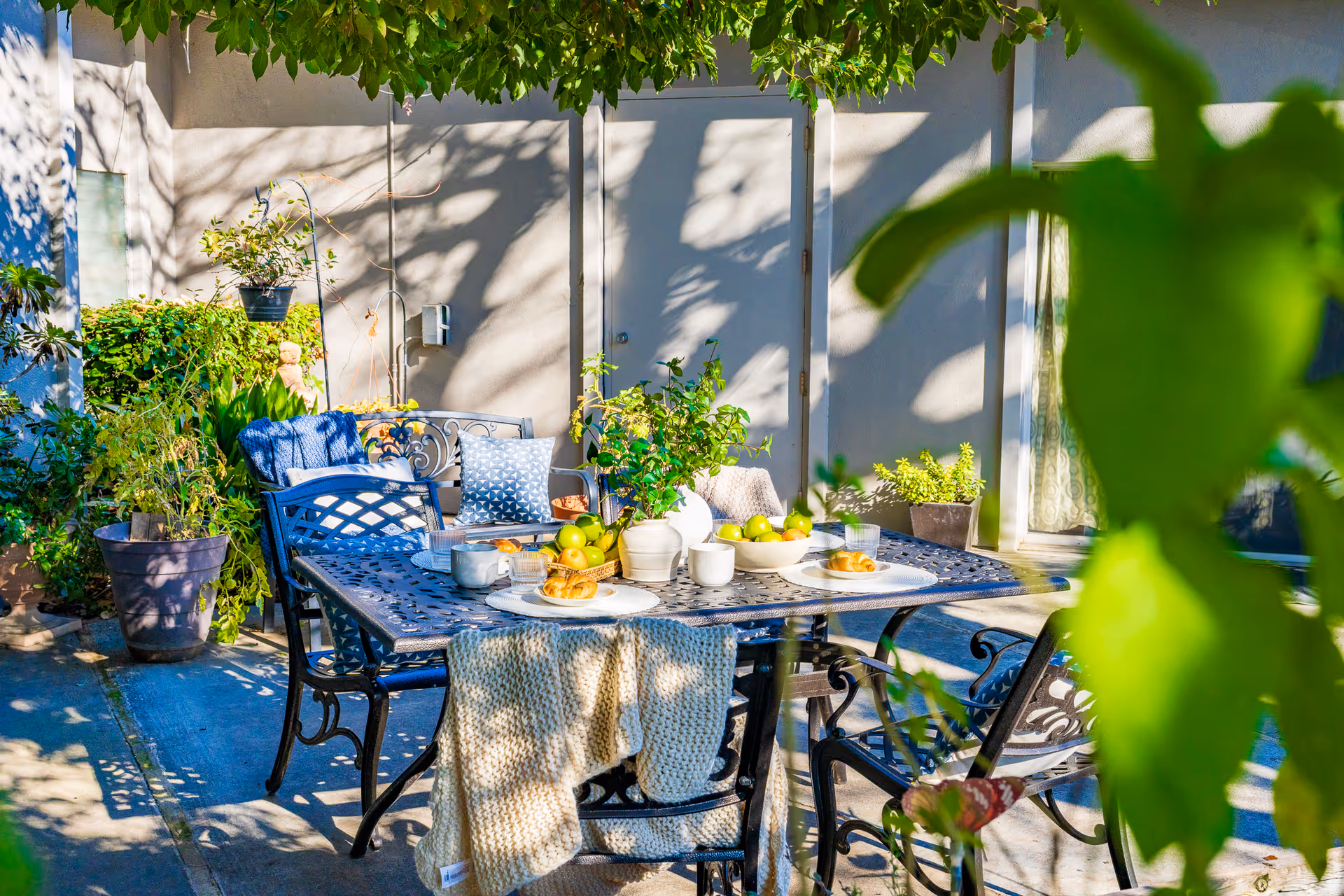 Outdoor patio area with a metal table and chairs set for breakfast, including plates with croissants, cups, a bowl of green apples, and a vase with green plants. The area is surrounded by potted plants and greenery, with sunlight casting shadows on the wall behind.