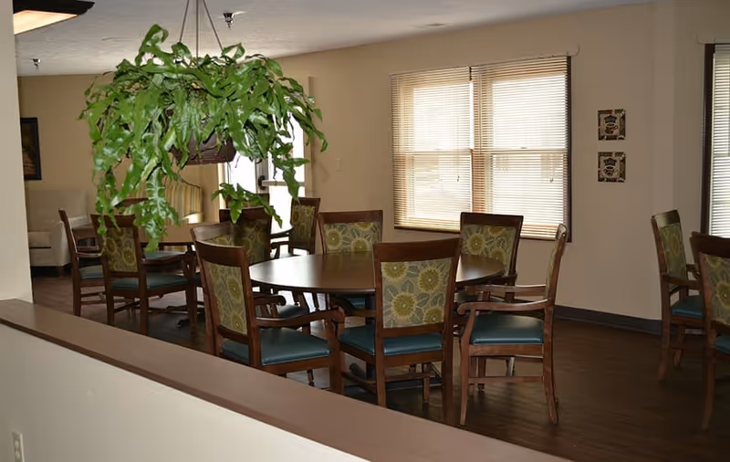 Interior view of a dining area with round wooden tables and chairs featuring floral patterned backs and green seats. A large hanging green plant is visible above one table. The room has beige walls, wooden flooring, and windows with blinds letting in natural light.