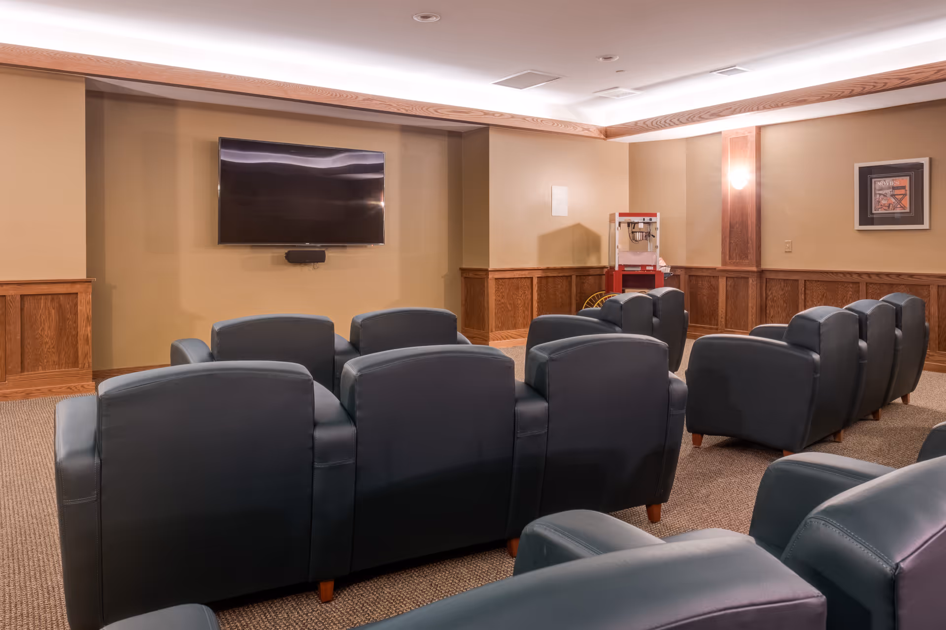 A cozy media room with multiple rows of dark leather recliner chairs facing a large flat-screen TV mounted on a beige wall. The room features wood paneling along the lower half of the walls, a popcorn machine in the corner, and a framed movie-themed picture on the wall.