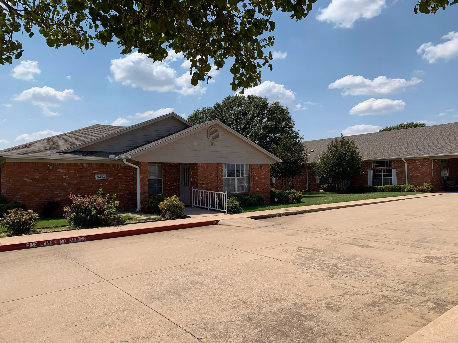 Front exterior of a single-story brick senior living building with a driveway, landscaped shrubs, and a blue sky.