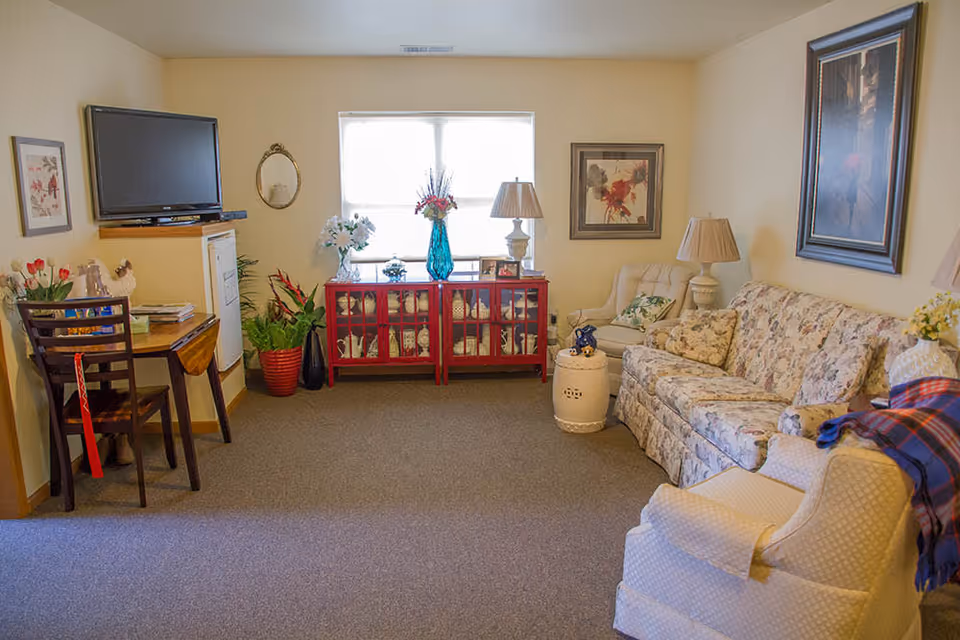 Cozy living room with a floral sofa and armchairs, a TV on the wall, a red display cabinet beneath a window, and lamps and plants.