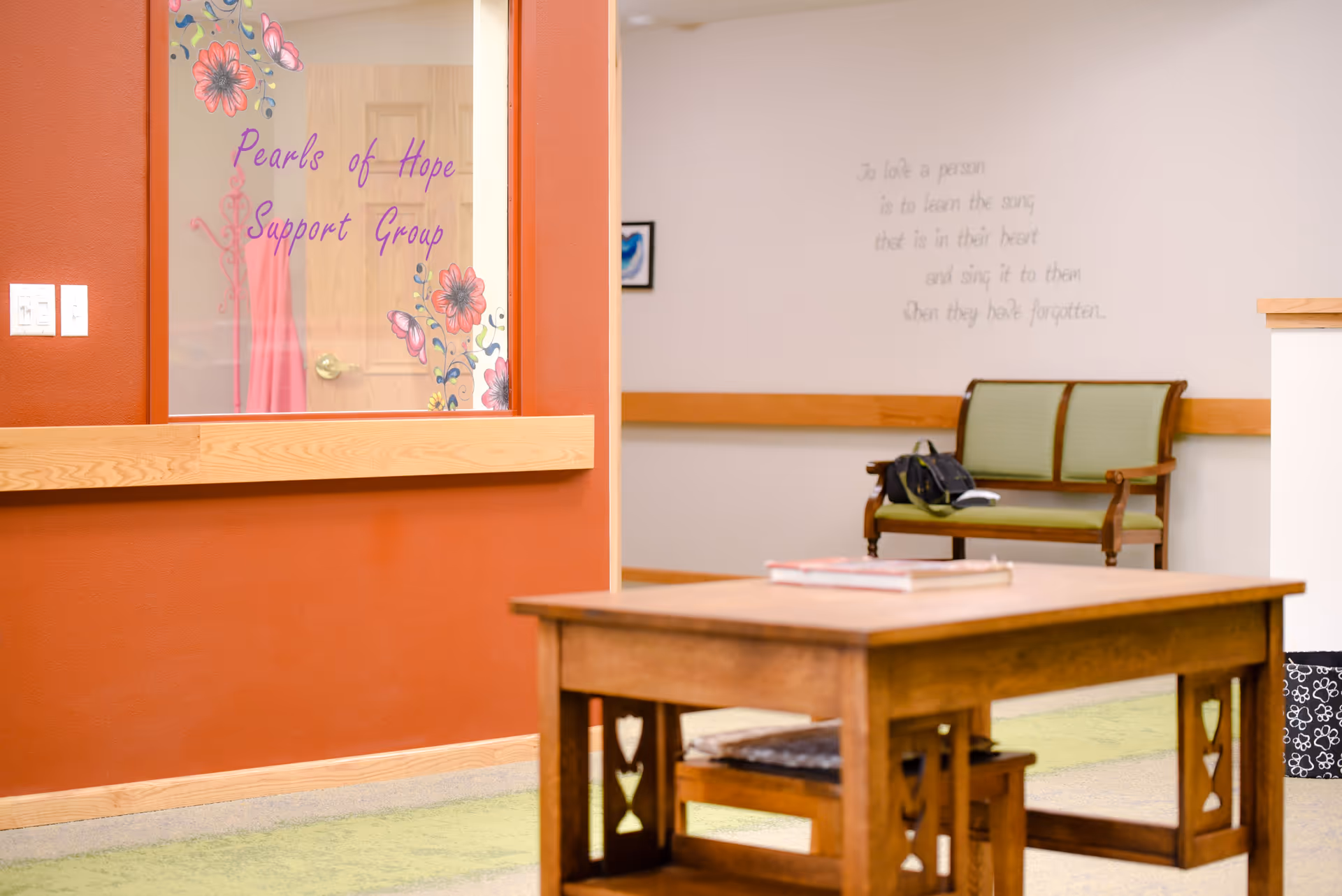 Interior common area with a wooden table in the foreground, a bench in the background and a glass door labeled "Pearls of Hope Support Group" on a red wall.