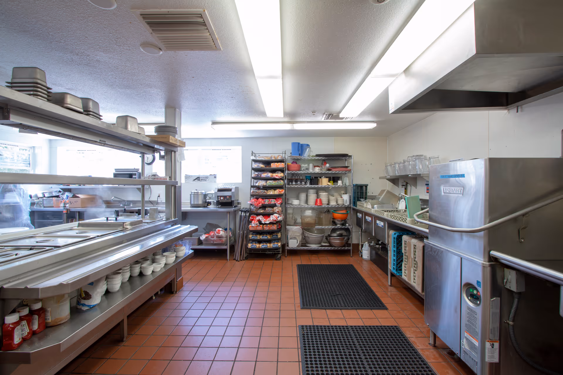 A clean and organized commercial kitchen with stainless steel counters, shelves stocked with plates, bowls, and food supplies, and industrial kitchen equipment including a dishwasher and food warming station. The floor is tiled with reddish-brown tiles and there are black anti-fatigue mats placed on the floor.