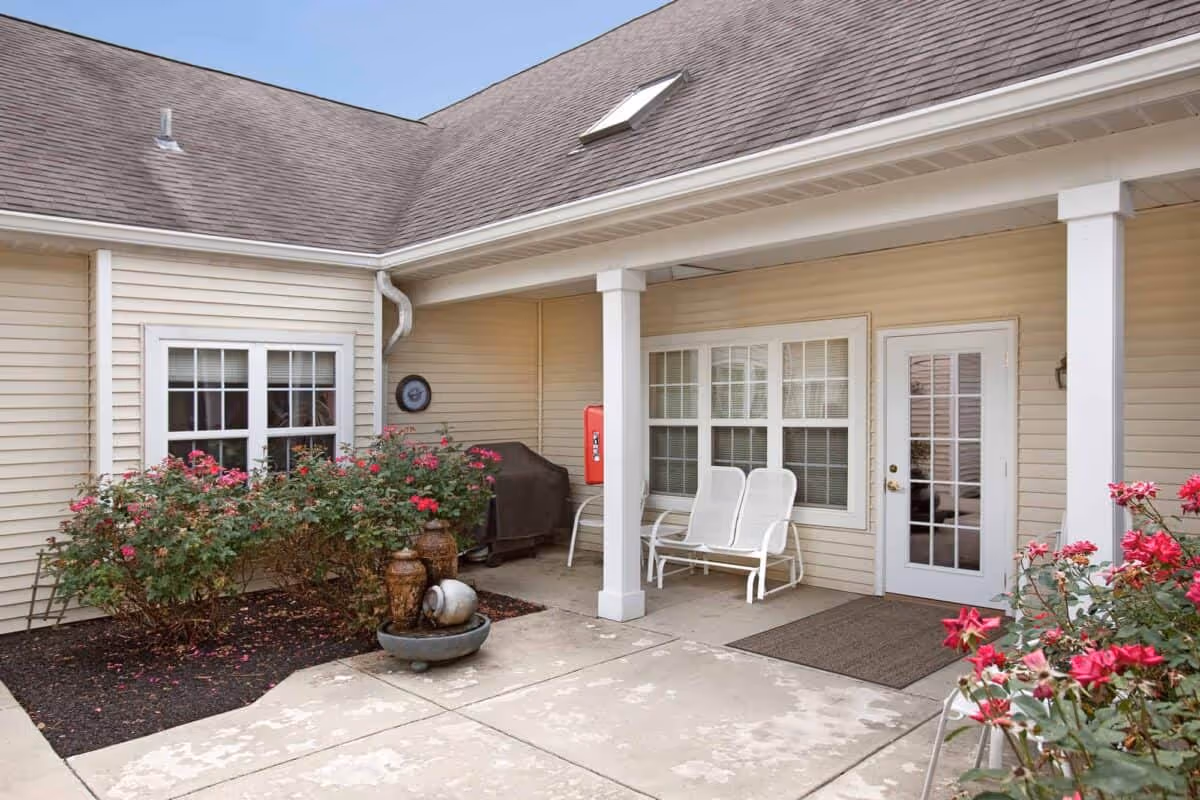 Covered exterior courtyard with white patio chairs, potted fountain, flowering shrubs, and a glass entry door on a beige-sided building.