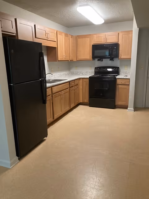 Kitchen with L-shaped wood cabinets, black refrigerator and stove, laminate countertops and linoleum floor.