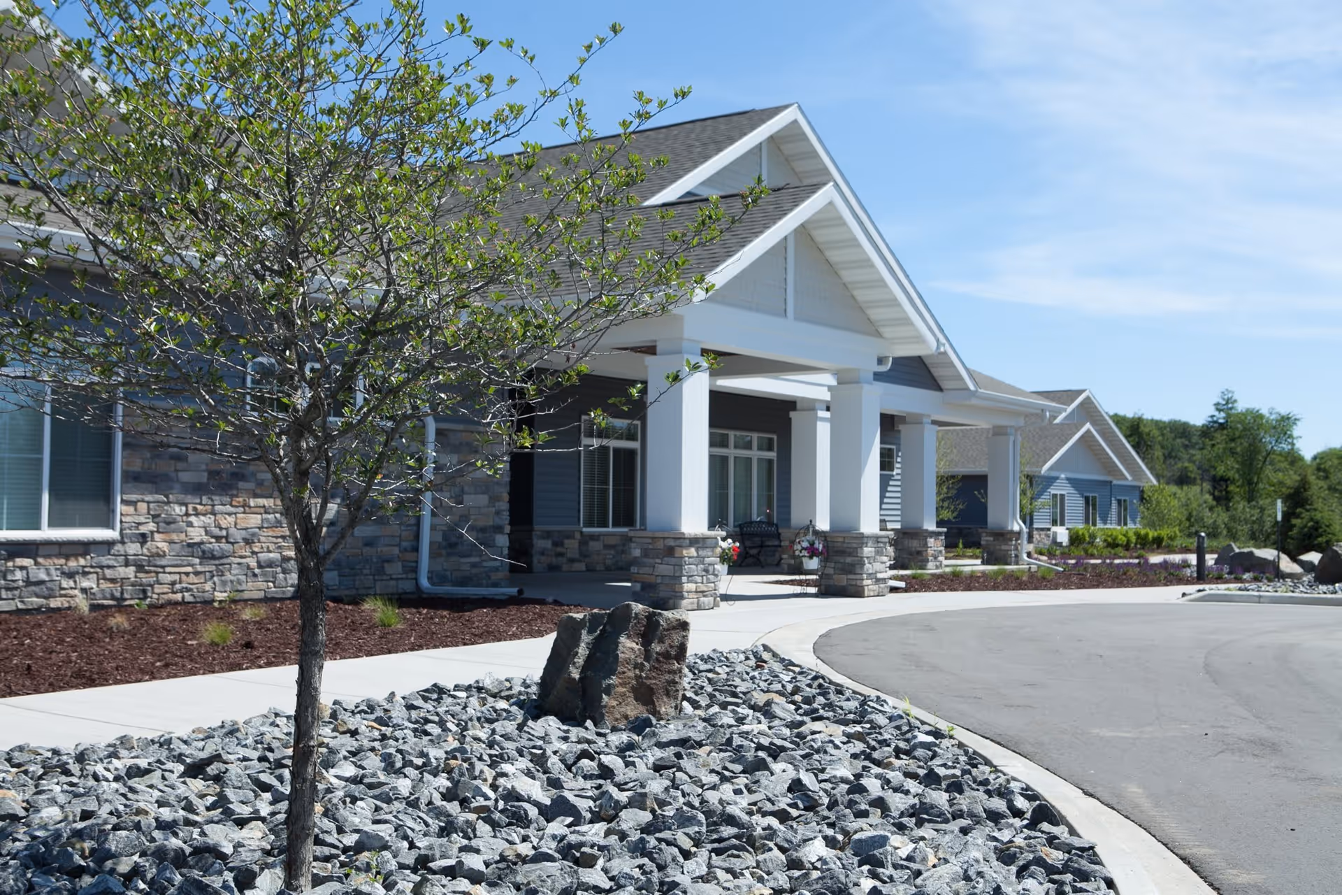 Exterior view of Frontida Assisted Living of Manitowoc showing a building with stone and blue siding, white pillars supporting a covered entrance, a small tree surrounded by rocks, and a curved driveway under a clear blue sky.