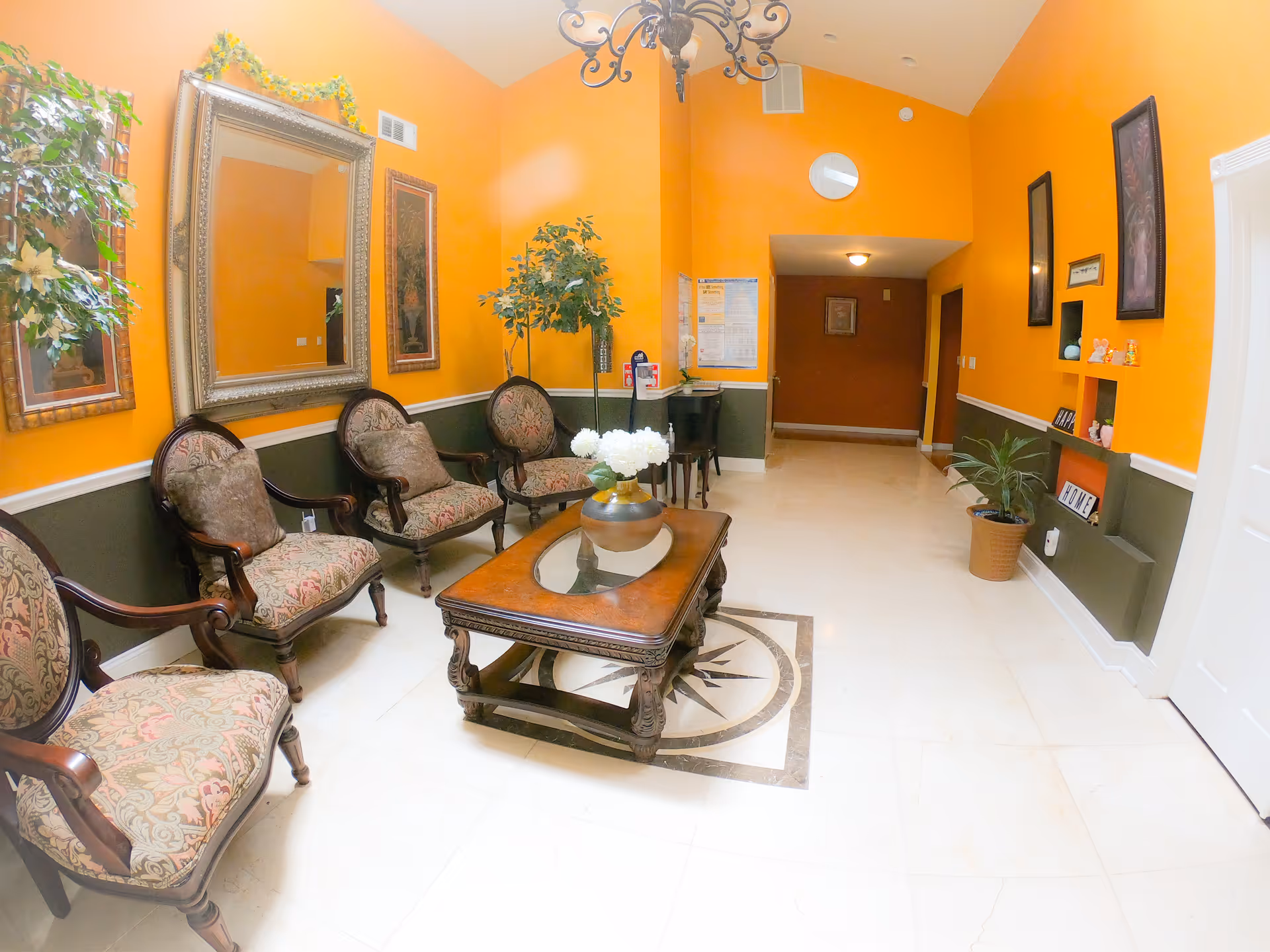 A cozy sitting area with four upholstered armchairs arranged around a wooden coffee table with a glass inset and a vase of white flowers. The walls are painted bright orange with a dark green wainscoting. There are framed artworks and a large decorative mirror on the walls. A chandelier hangs from the ceiling, and there are potted plants adding greenery to the space.