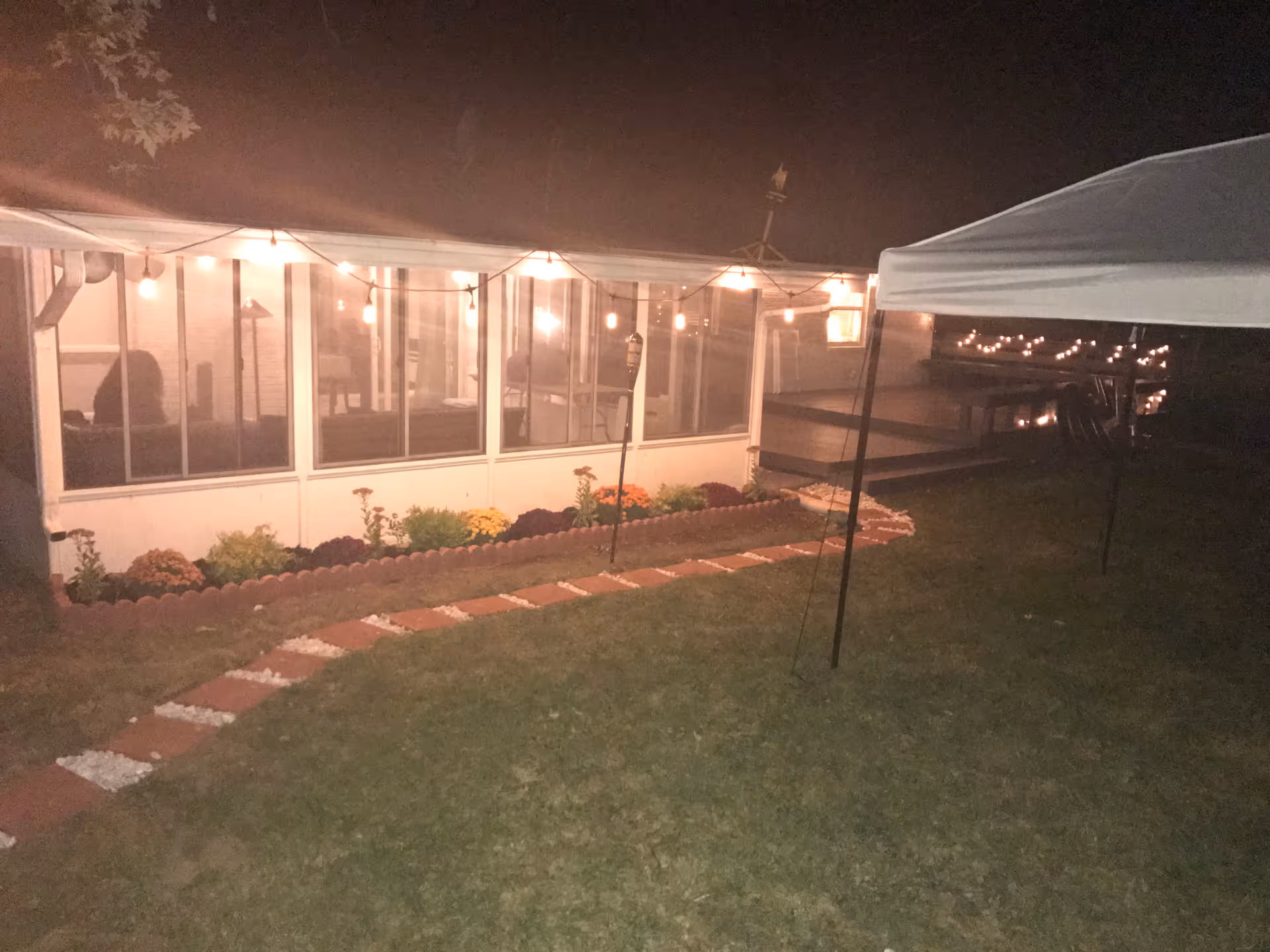 Nighttime view of a backyard garden area with a pathway made of red bricks and white stones leading to a porch with string lights hanging above. There are flowers planted along the edge of the porch, and a canopy tent is partially visible on the right side of the image.