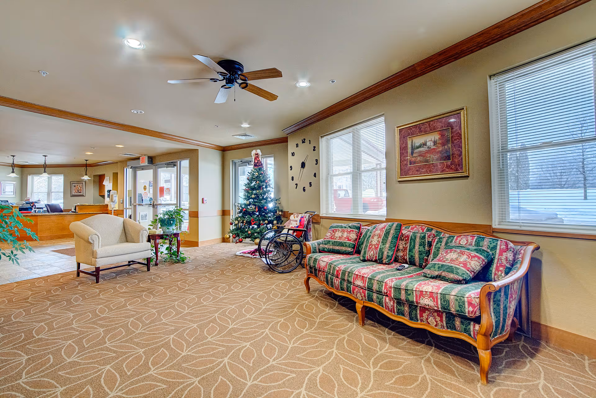 Interior view of a senior living facility lobby area with a patterned carpet, a Christmas tree decorated with ornaments, a wheelchair, a vintage-style sofa with green and red upholstery, a beige armchair, a small table with plants, large windows with blinds, a wall clock with black numbers, and framed artwork on the wall.