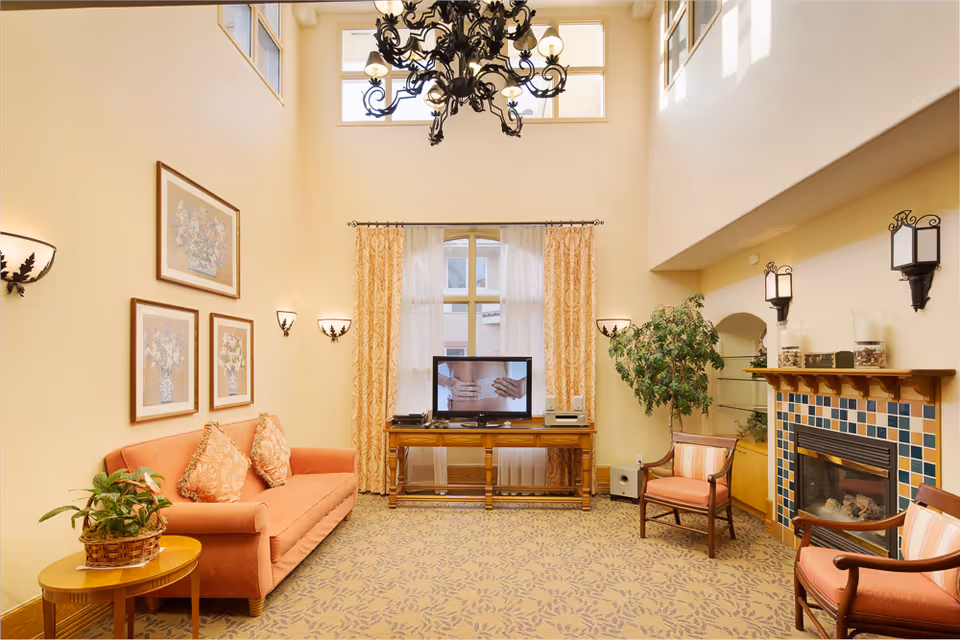 High-ceilinged living room with a coral sofa, TV on a wooden console, a tiled fireplace, chandelier, and additional seating and plants.