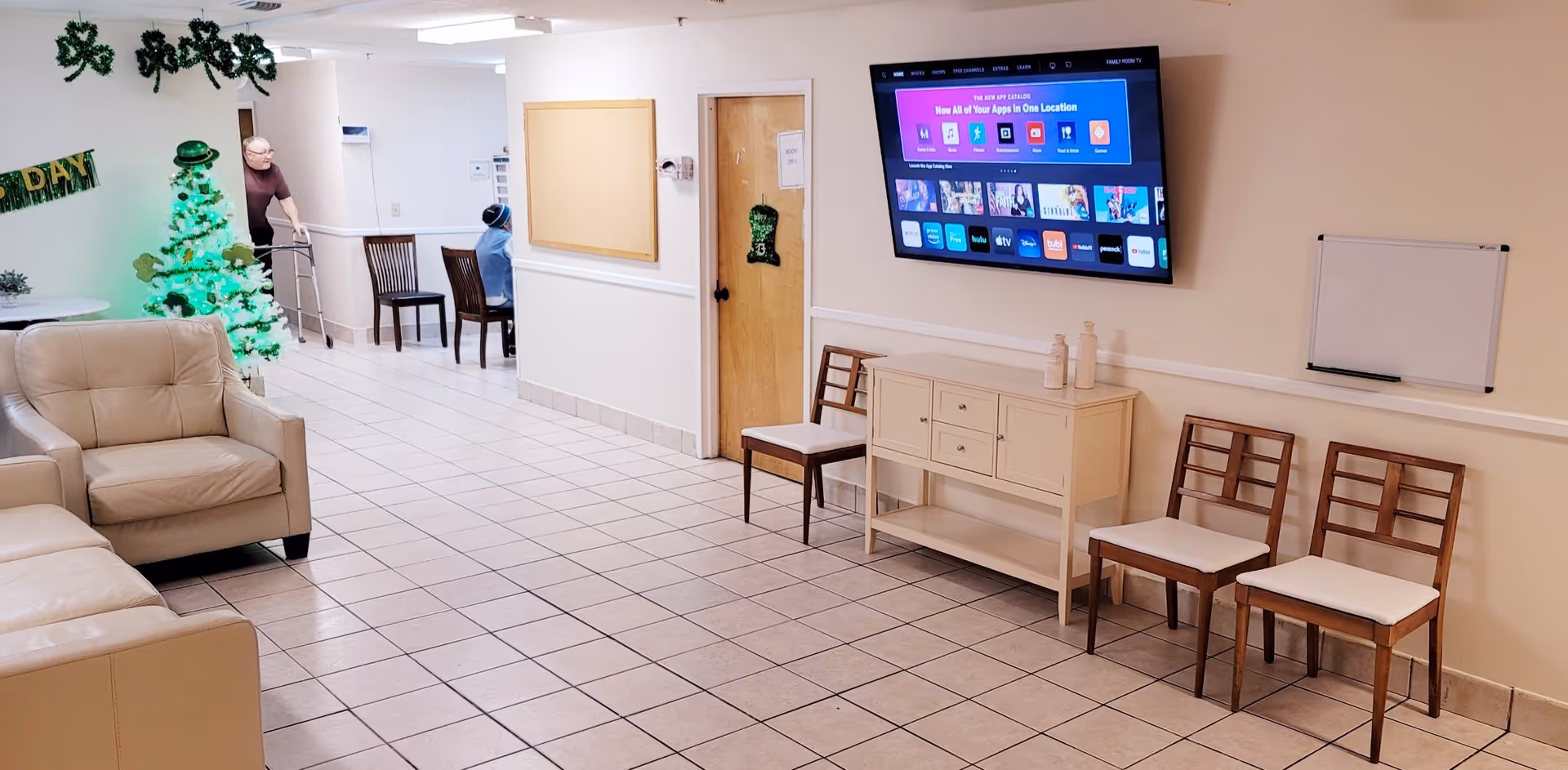 A hallway in a senior living facility decorated for St. Patrick's Day with a green shamrock garland and a green-lit Christmas tree. There are beige leather chairs on the left, wooden chairs along the right wall, a wall-mounted TV displaying a smart TV interface, and a small cabinet beneath it. Two elderly people are visible in the background, one using a walker.