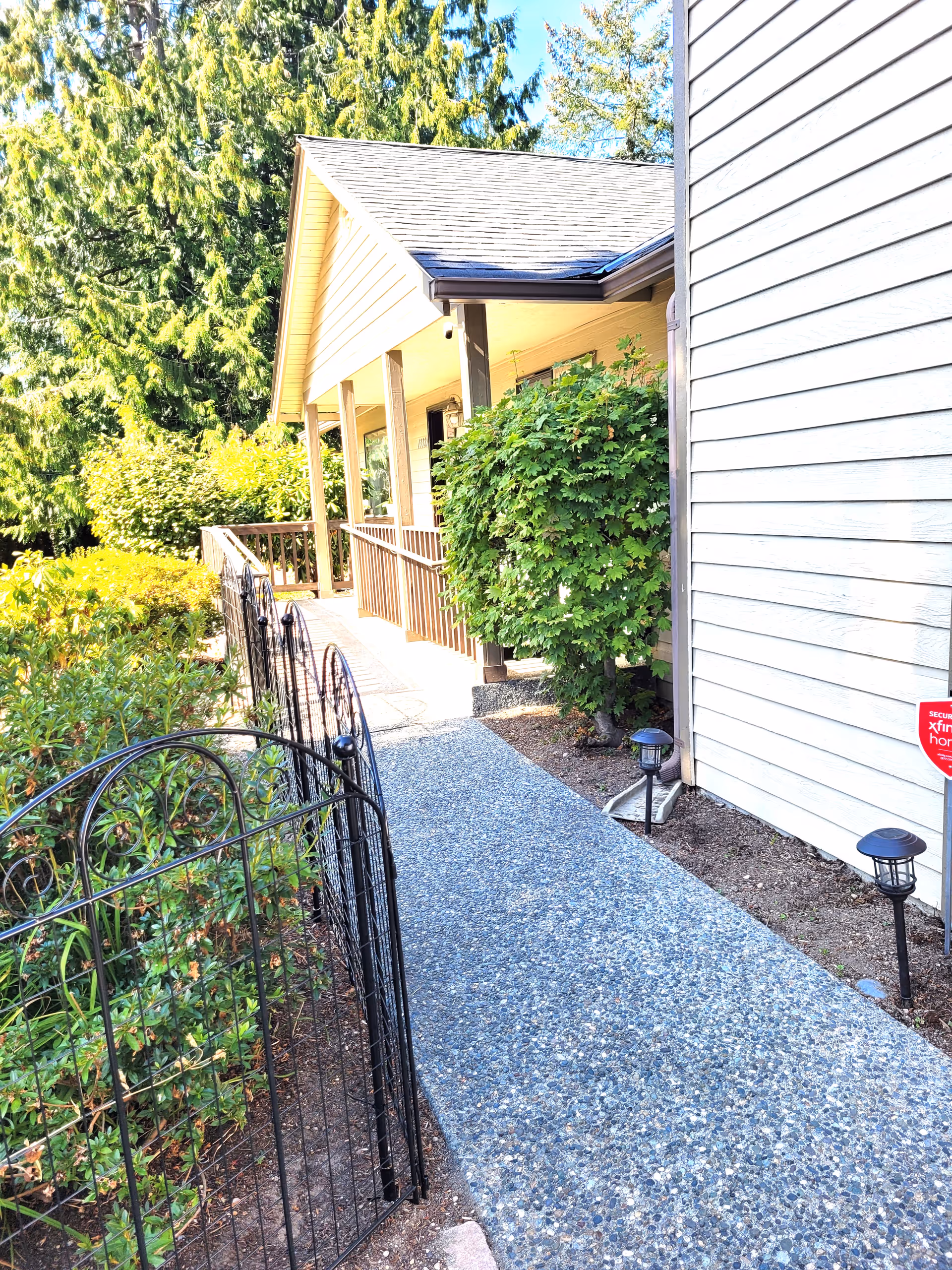 A paved walkway leading to the entrance of a house with a covered porch supported by wooden pillars. The house exterior is light-colored with horizontal siding. There is a black metal fence on the left side of the walkway and green bushes and trees surrounding the area. Two small garden lights are visible along the right side of the walkway, and a red security sign is posted near the house wall.