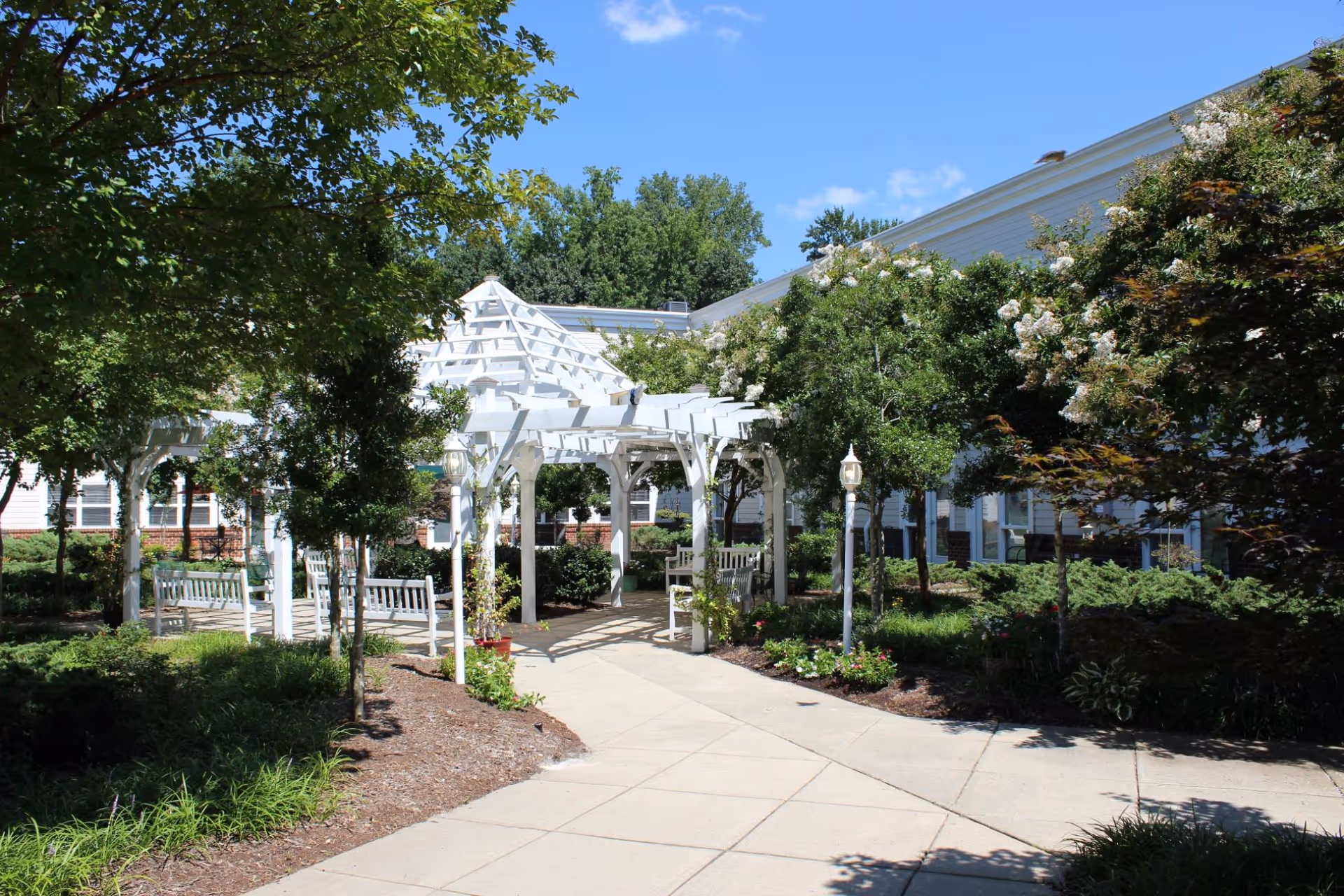 A sunny outdoor garden area with a white wooden pergola surrounded by green trees and shrubs. There are benches under the pergola and a paved walkway leading to it. The building exterior with white siding and windows is visible in the background under a clear blue sky.