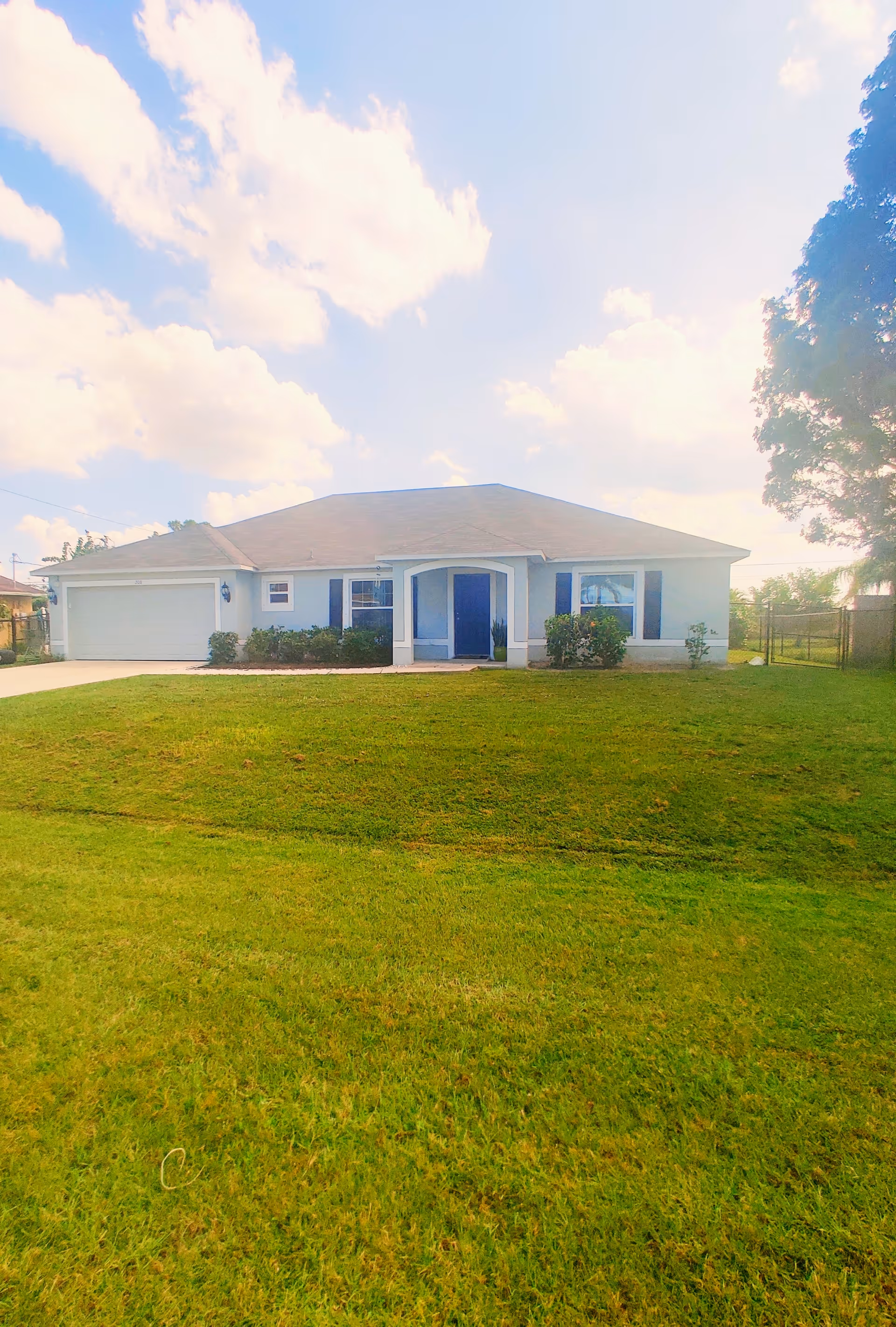 Single-story suburban house with a wide green front lawn, central covered entry, attached garage, and blue shutters under a partly cloudy sky.