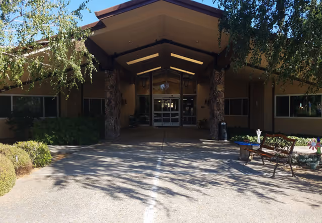 Front entrance of a building with a covered walkway supported by stone pillars, surrounded by bushes and trees casting shadows on the concrete path. There is a bench and some potted plants to the right side near the entrance.