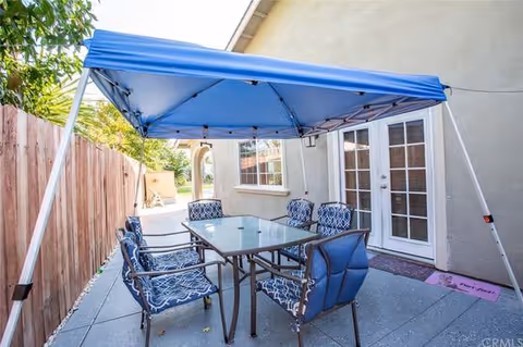 Outdoor patio area with a blue canopy tent covering a glass-top table surrounded by six cushioned chairs with blue patterned fabric. The patio is adjacent to a beige building with a window and double glass doors. A wooden fence runs along one side of the patio.