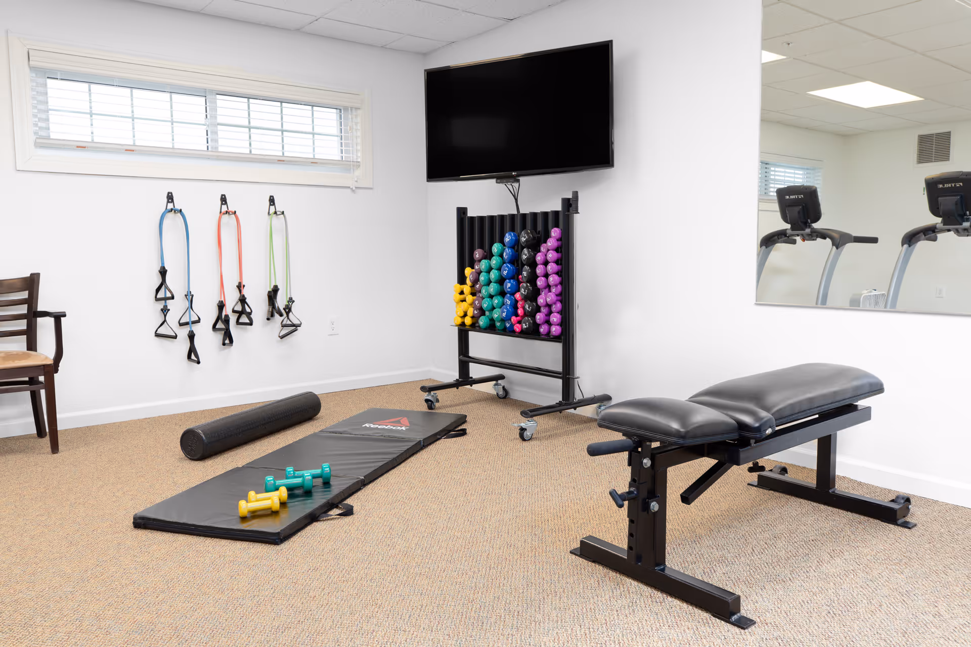 Small fitness room with an exercise bench, mat, foam roller, resistance bands, colorful dumbbells, and a wall-mounted TV.