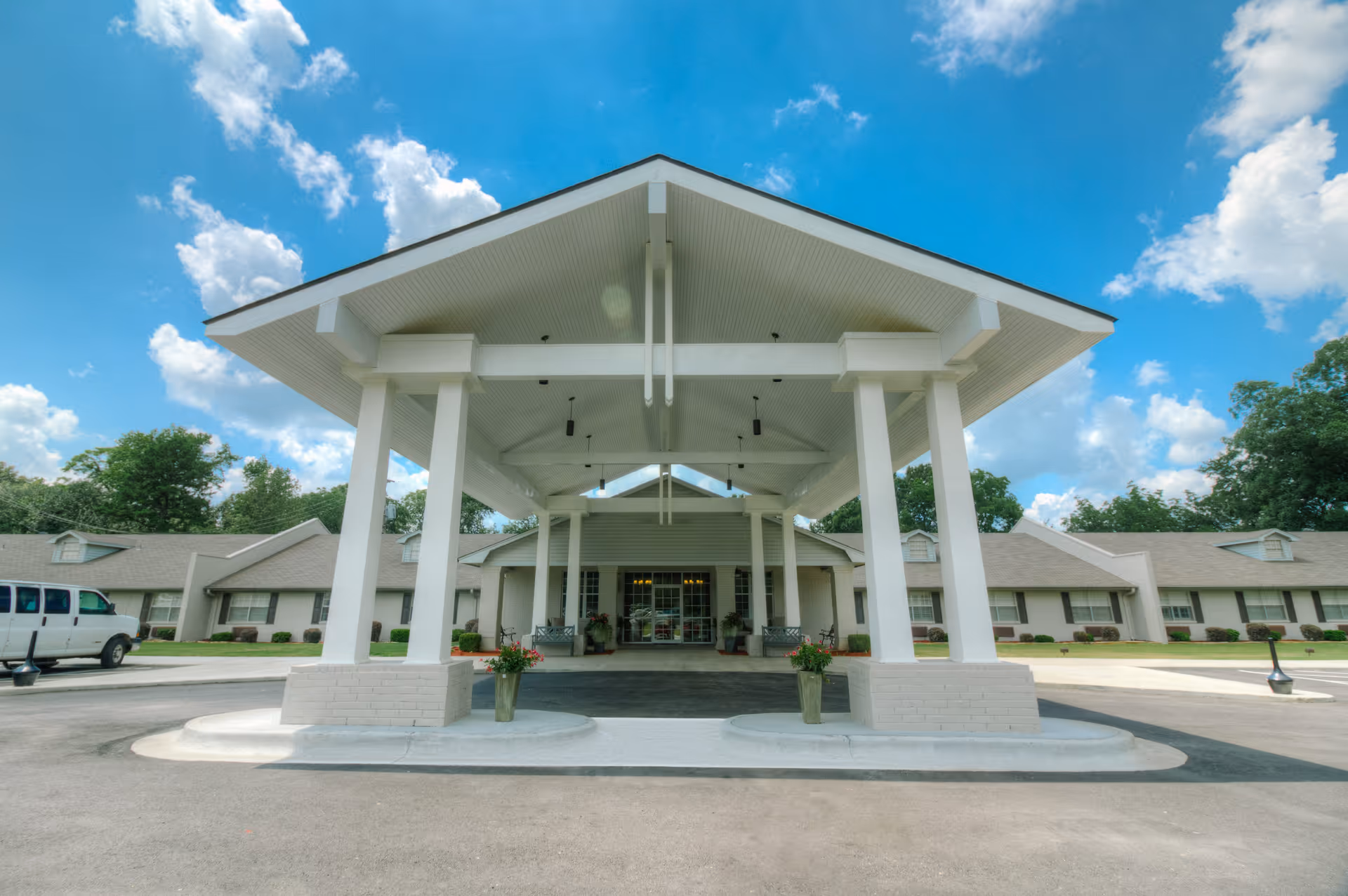Front exterior view of Spring Creek Health & Rehab facility showing a large covered entrance with white pillars, a driveway, and a building with multiple windows under a blue sky with clouds.