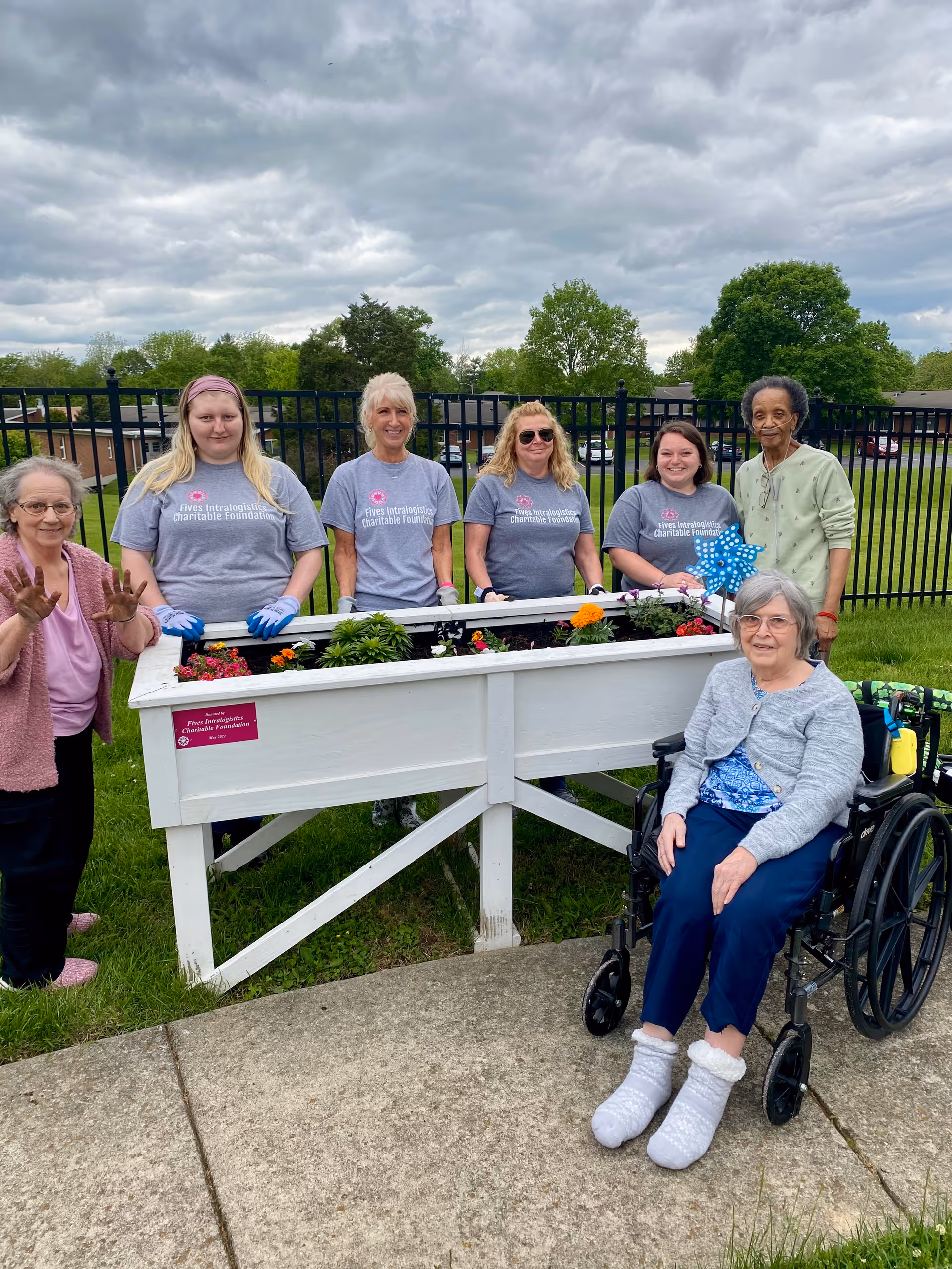 A group of six women, including one in a wheelchair, standing and sitting around a raised garden bed filled with flowers outdoors. Four women are wearing matching gray t-shirts with a charitable foundation logo. The background shows a black metal fence, green grass, trees, and a cloudy sky.