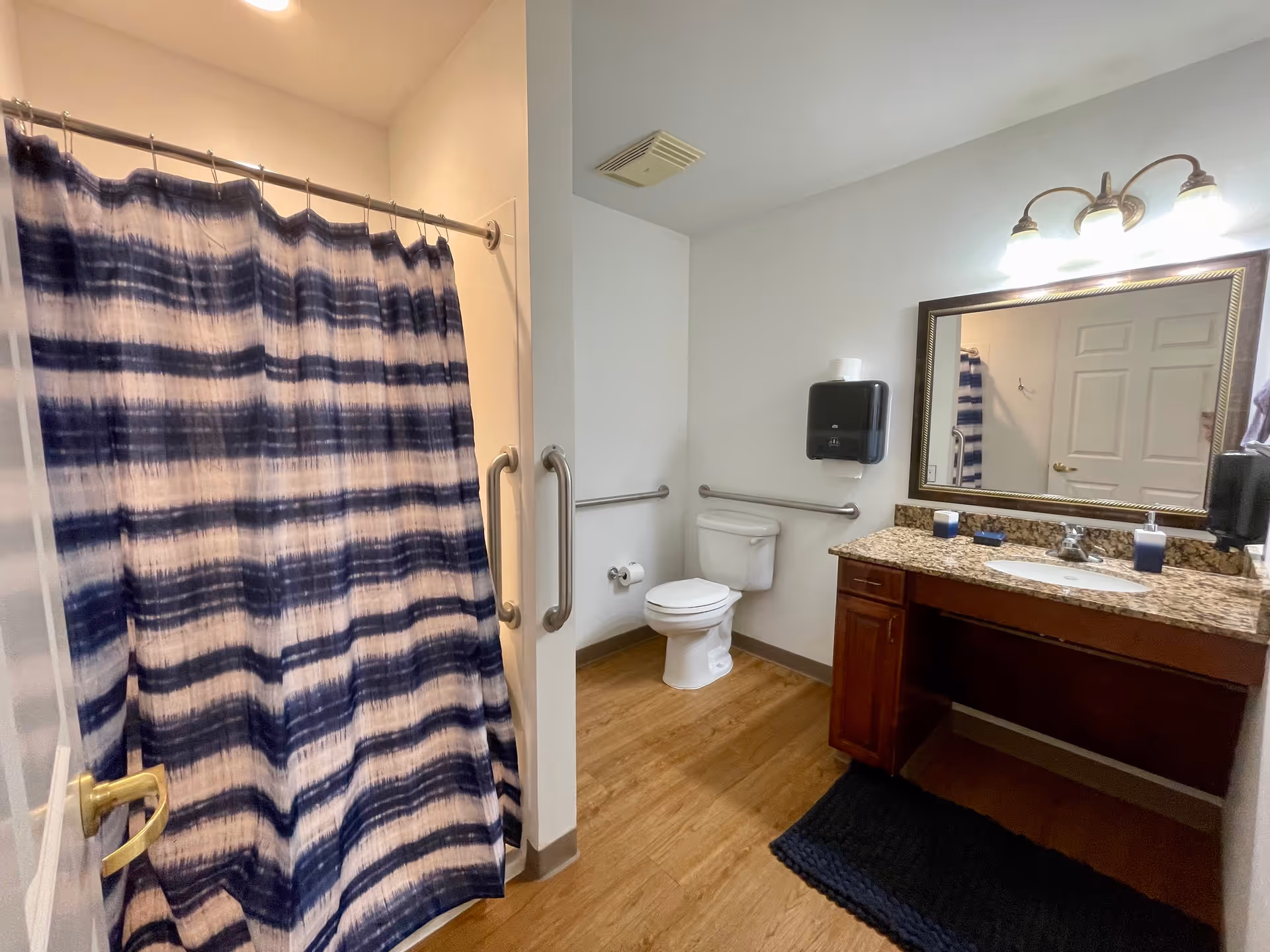 Bathroom with a shower area enclosed by a blue and white striped curtain, a toilet with grab bars on the walls, a wooden vanity with a granite countertop, a sink, a large mirror above the sink, and a black paper towel dispenser mounted on the wall.