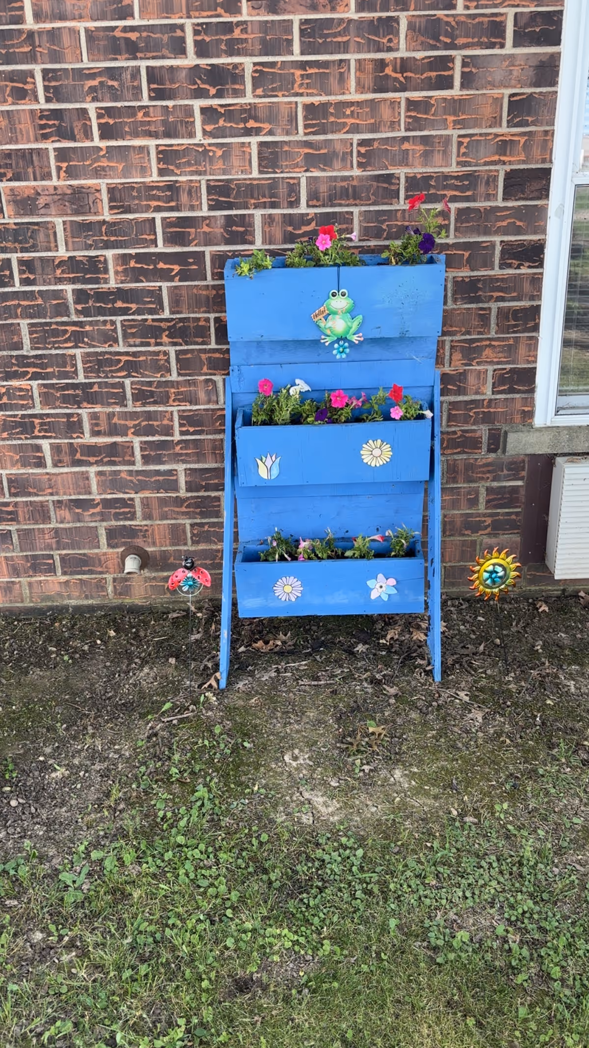 Blue tiered wooden planter filled with flowers and decorative ornaments against a brick exterior wall.