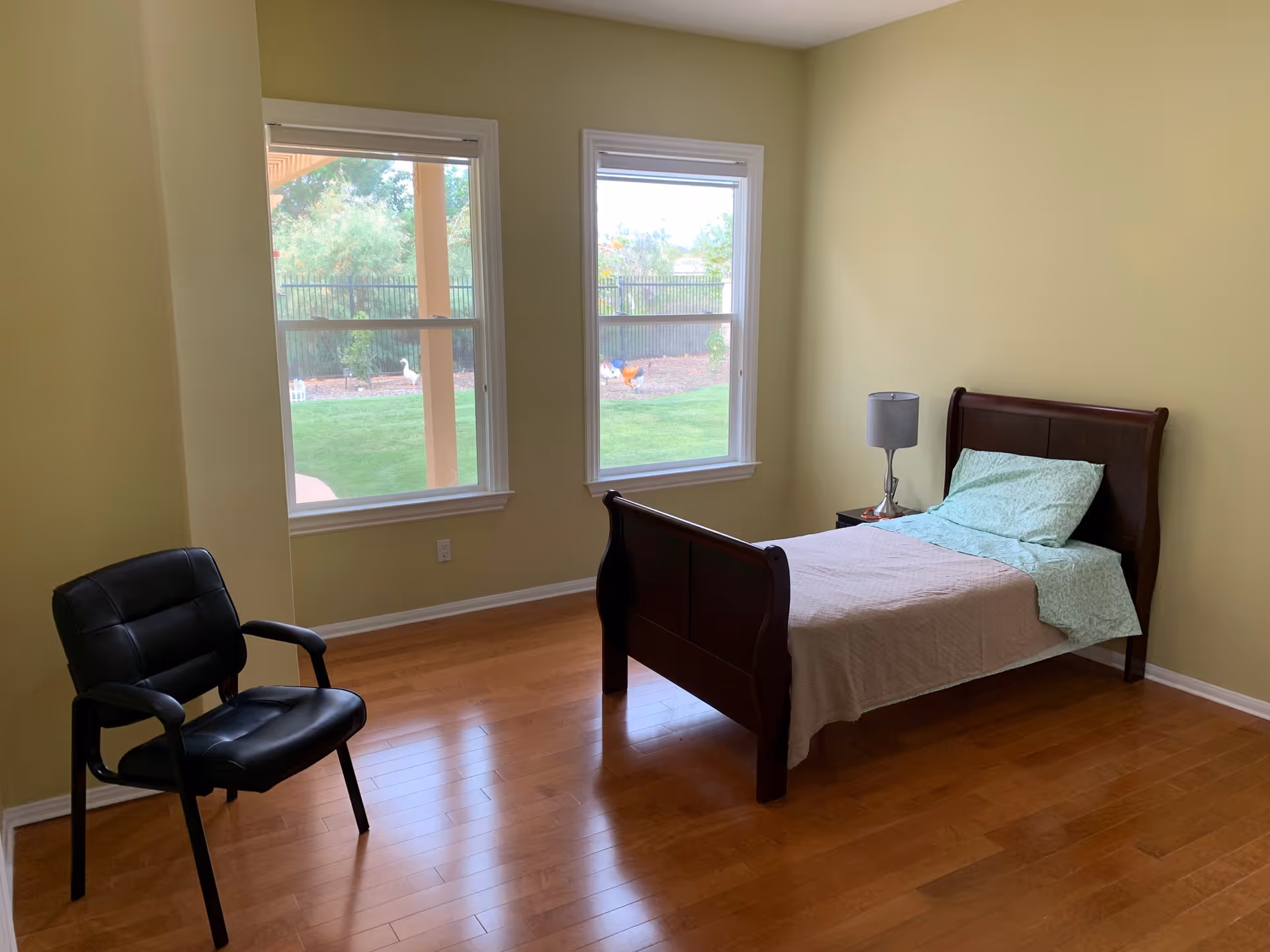 Sparsely furnished bedroom with a single bed, nightstand and chair beside two large windows overlooking a yard.