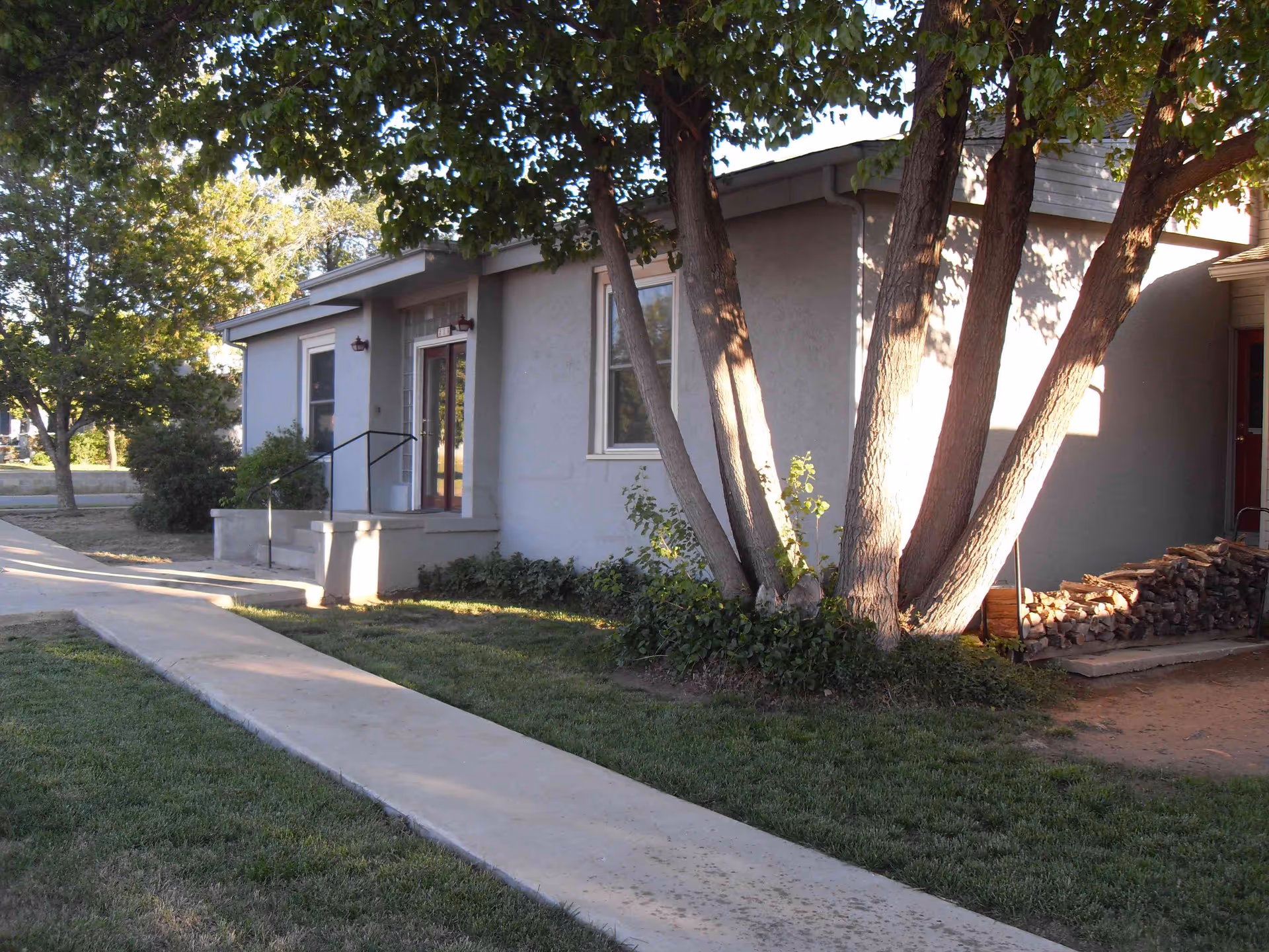 Exterior view of a single-story building with a light gray facade, a small set of stairs leading to a door with glass panels, surrounded by green grass and trees providing shade. A pile of firewood is stacked near the building on the right side.
