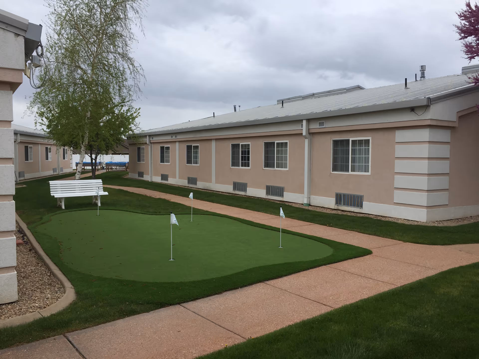Courtyard featuring a small putting green with flags, a white bench and walkways in front of single-story beige buildings under a cloudy sky.