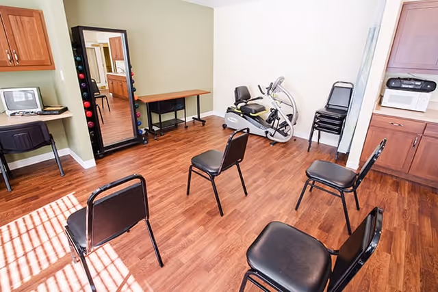 Small senior living activity/fitness room with chairs arranged in a circle, an exercise machine, a mirror with hand weights, and a desk with a computer.