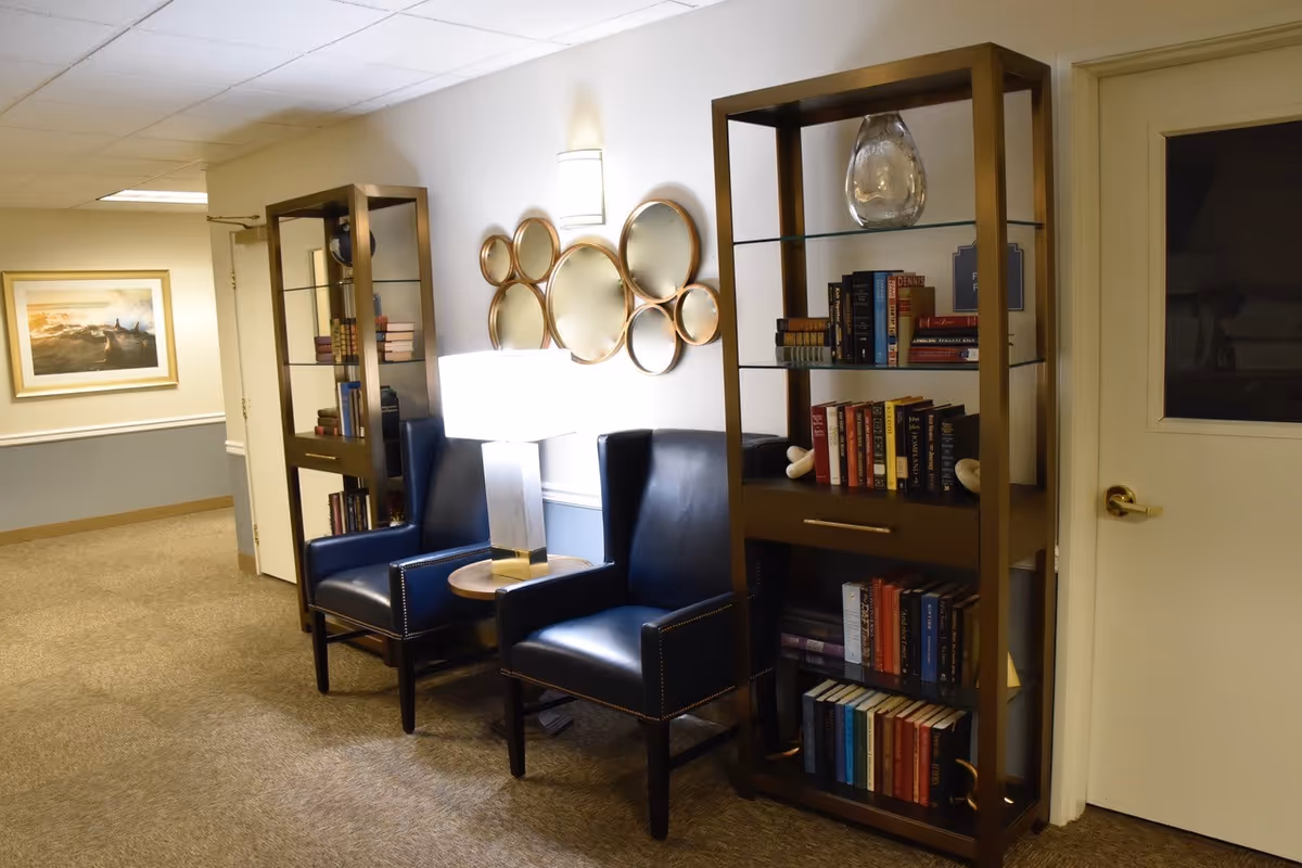 Two navy armchairs with a table lamp between tall bookshelves and decorative round mirrors in a carpeted seating area.