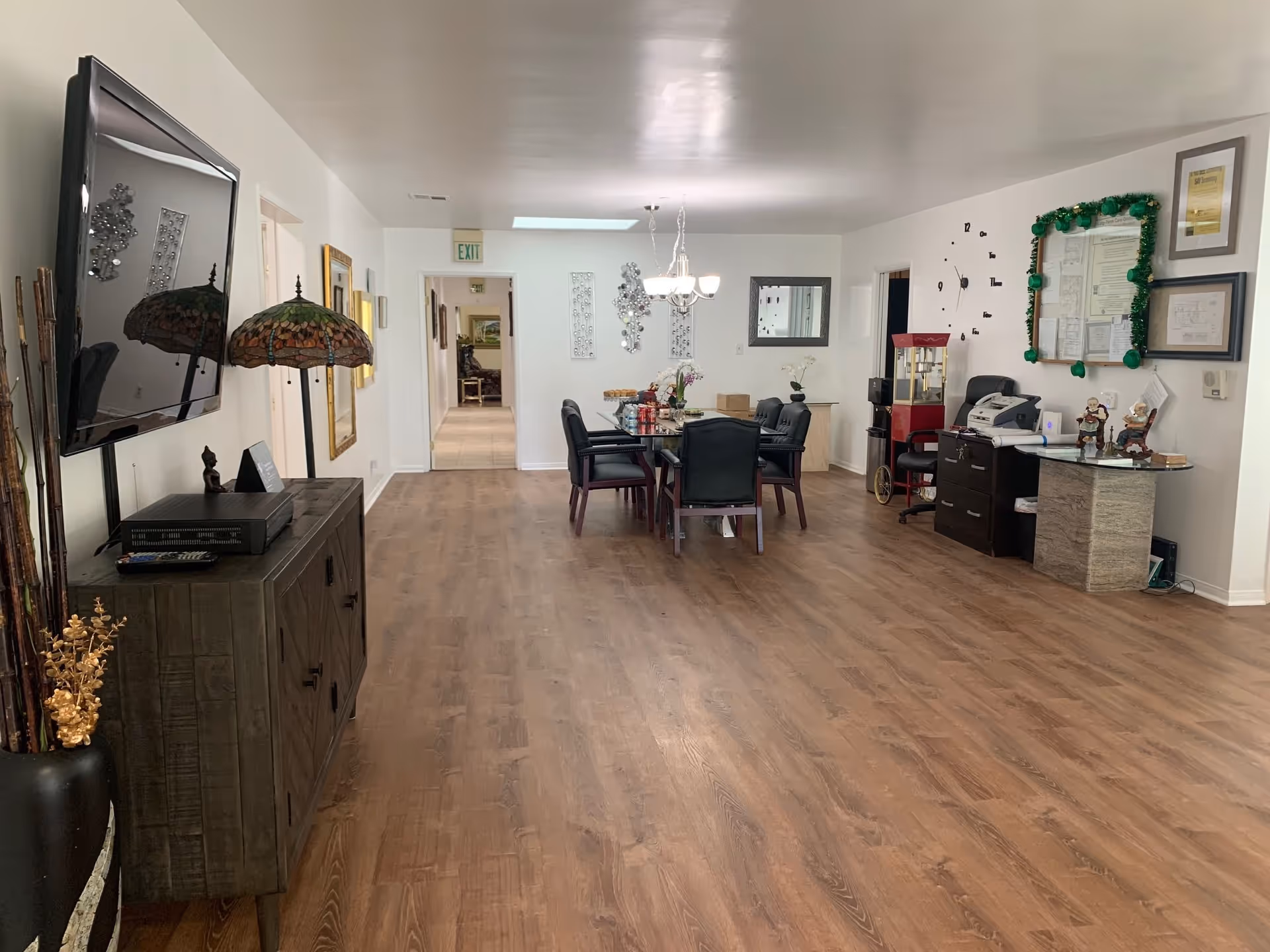 Interior view of a senior living facility room with wooden flooring, a wall-mounted TV on the left, a decorative lamp, and a dining table with six chairs in the center. The back wall has a chandelier, wall art, and a mirror. On the right side, there is a small office area with a desk, chair, a popcorn machine, and a bulletin board decorated with green garland. The room is well-lit with white walls and ceiling.