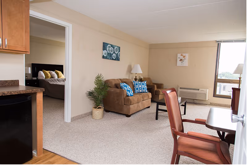 Interior view of a senior living facility apartment showing a living area with two brown armchairs adorned with blue and white patterned pillows, a small black coffee table, a lamp on a side table, and a potted plant. To the left, there is a partial view of a kitchen area with wooden cabinets and a black mini-fridge. In the background, a bedroom with a bed and pillows is visible through an open doorway. A window on the right side lets in natural light.