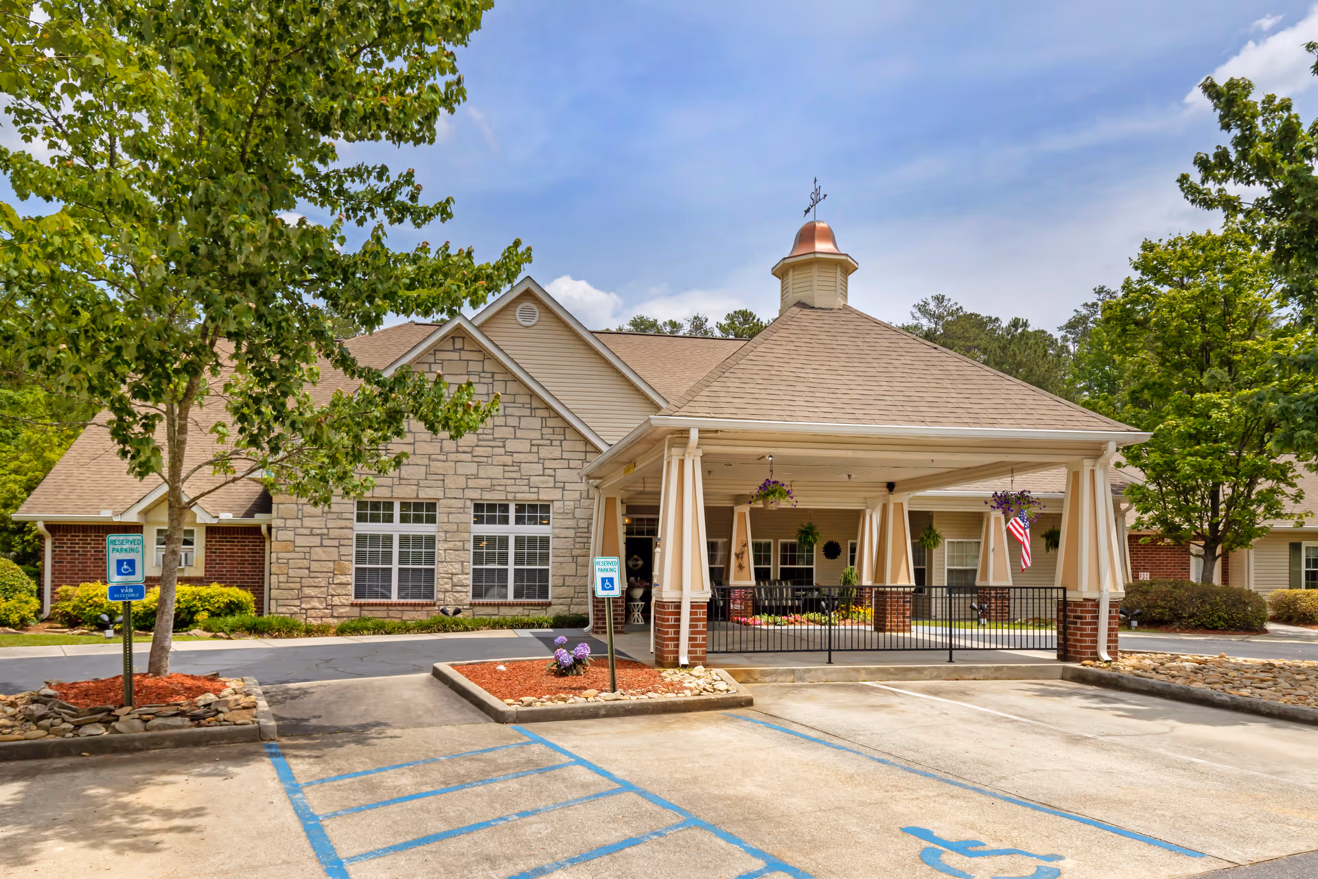 Exterior view of Brookdale Harbison facility showing a covered entrance with columns and a small cupola on the roof. There are two reserved handicapped parking spaces in front, landscaped areas with trees and flowers, and a clear blue sky above.