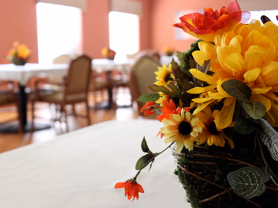 Close-up of a floral centerpiece with yellow and orange flowers on a table in a dining room with chairs and tables in the background.
