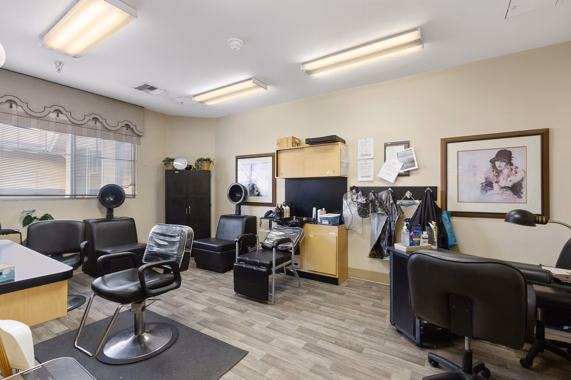 Interior view of a hair salon room in Claremont Place featuring black salon chairs, hair drying stations, a cabinet with hair products, and a framed artwork on the wall. The room has wood-patterned flooring and fluorescent ceiling lights.