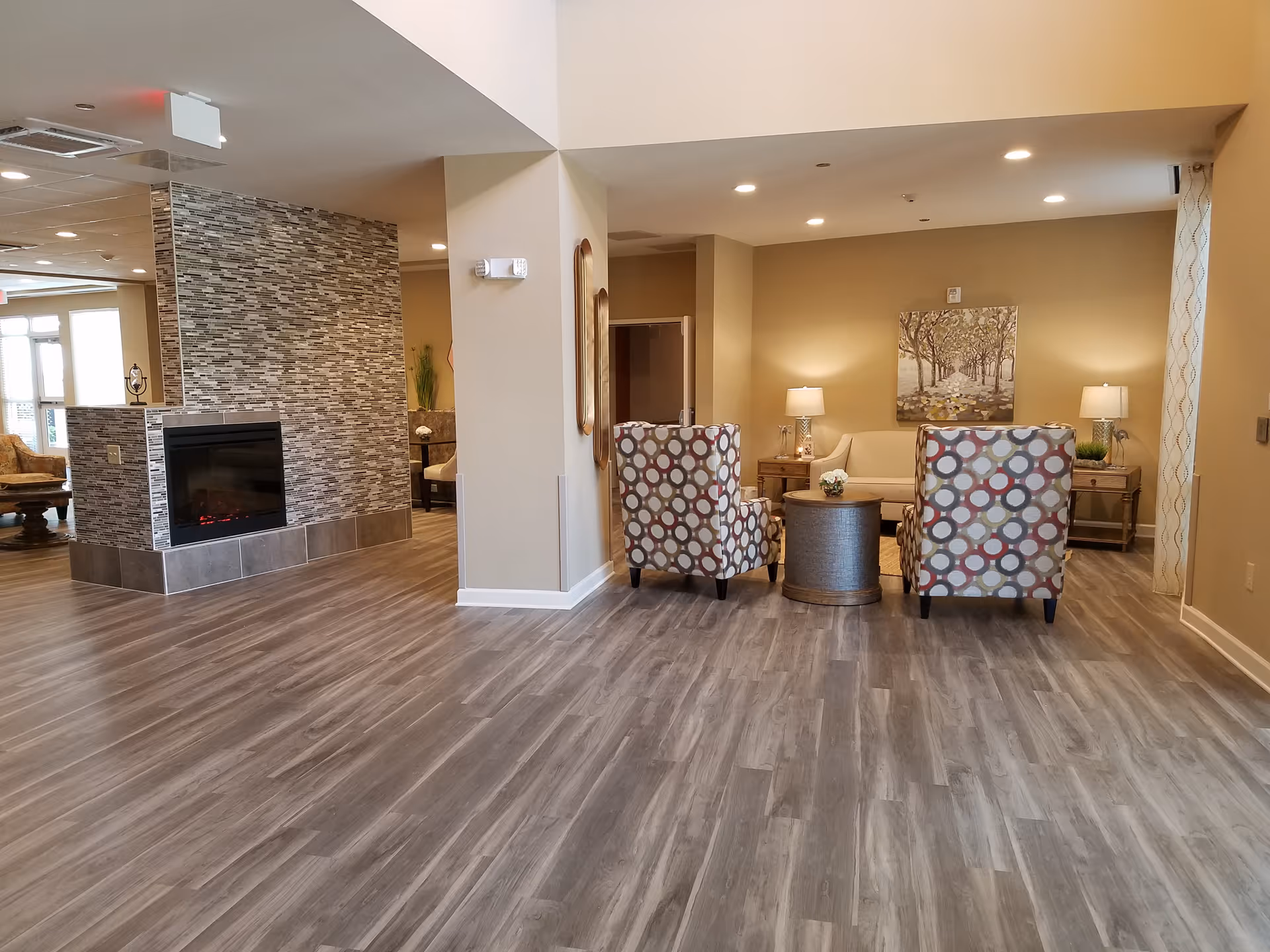 A cozy living room area with two patterned armchairs and a beige sofa arranged around a small round table. The room features wood-look flooring, warm beige walls, two side tables with lamps, a large painting of trees on the wall, and a modern fireplace with a tiled surround. The space is well-lit with recessed ceiling lights.