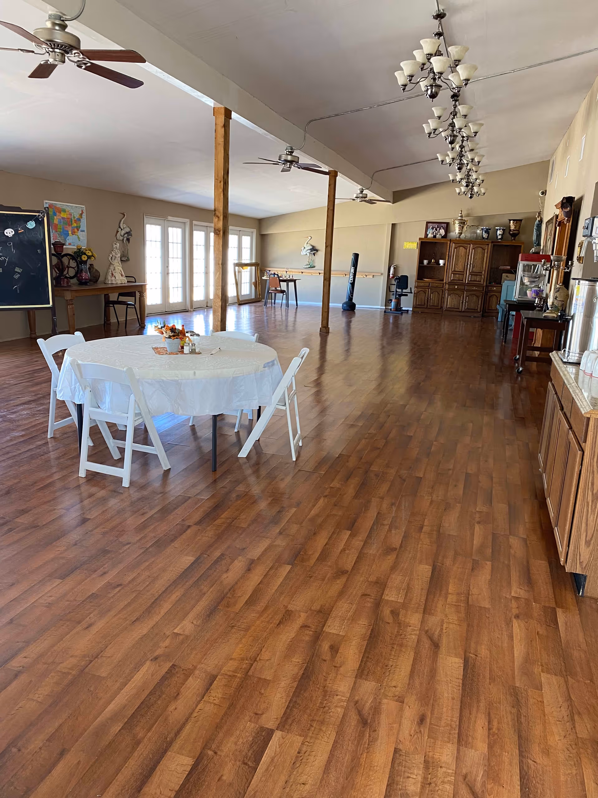 Spacious multipurpose dining/activity room with a round table and folding chairs, wood floors, ceiling fans, chandeliers, and cabinets along the walls.
