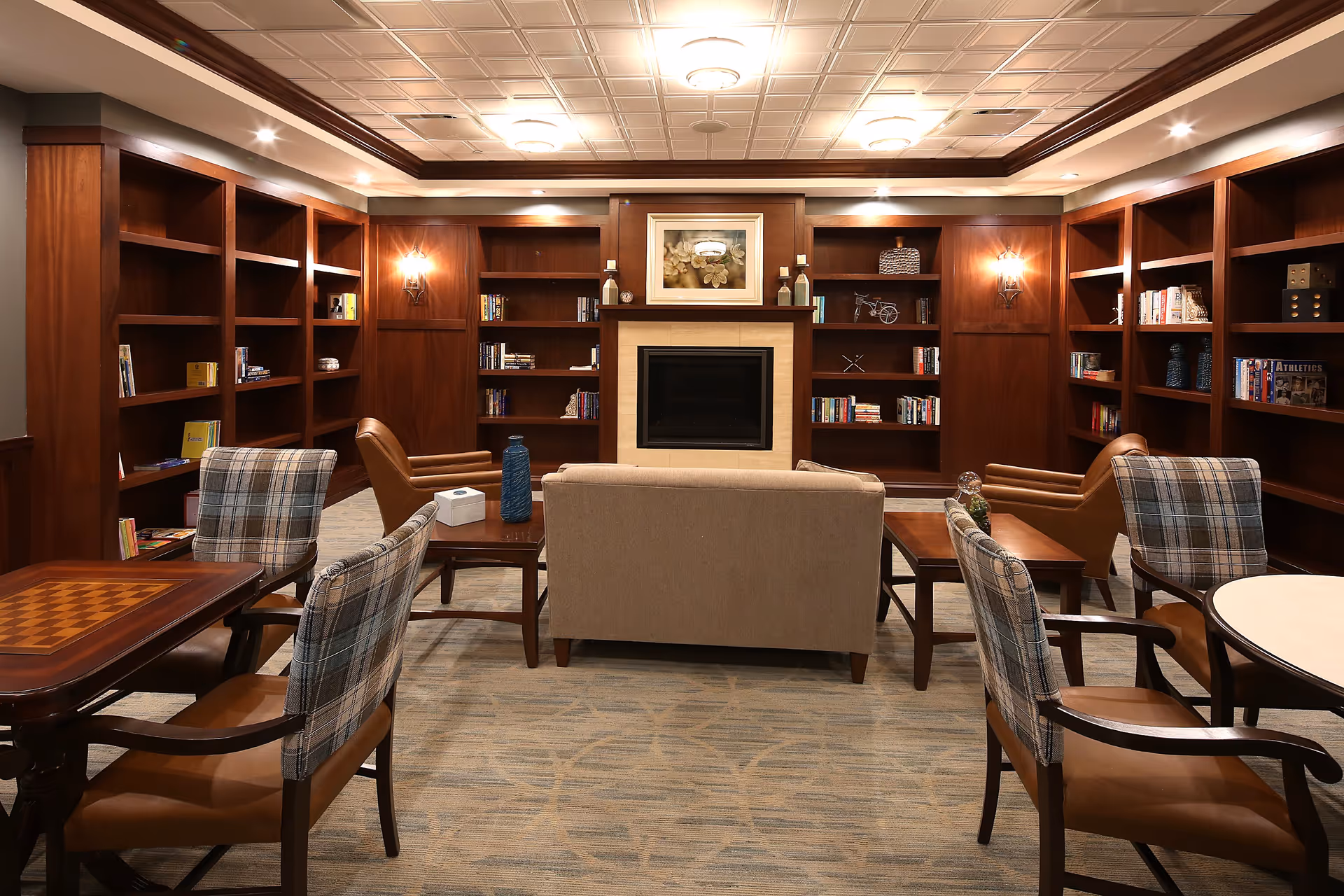 Cozy common room with built-in wood bookshelves, a central fireplace, sofas and armchairs around tables.