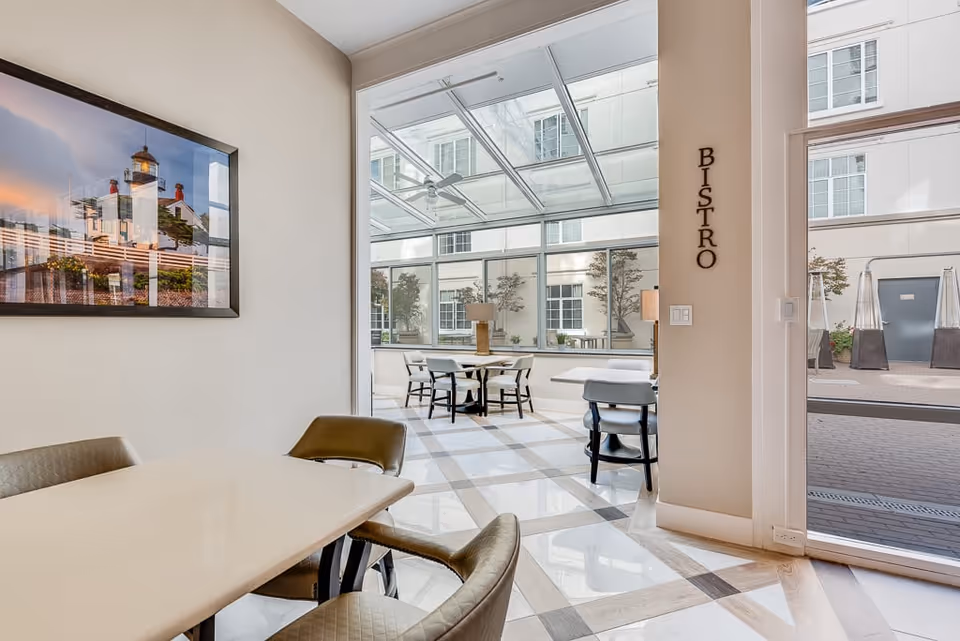 Interior view of a bright dining area labeled 'Bistro' with modern tables and chairs, large windows, and a glass ceiling allowing natural light. A framed picture of a lighthouse hangs on the wall.