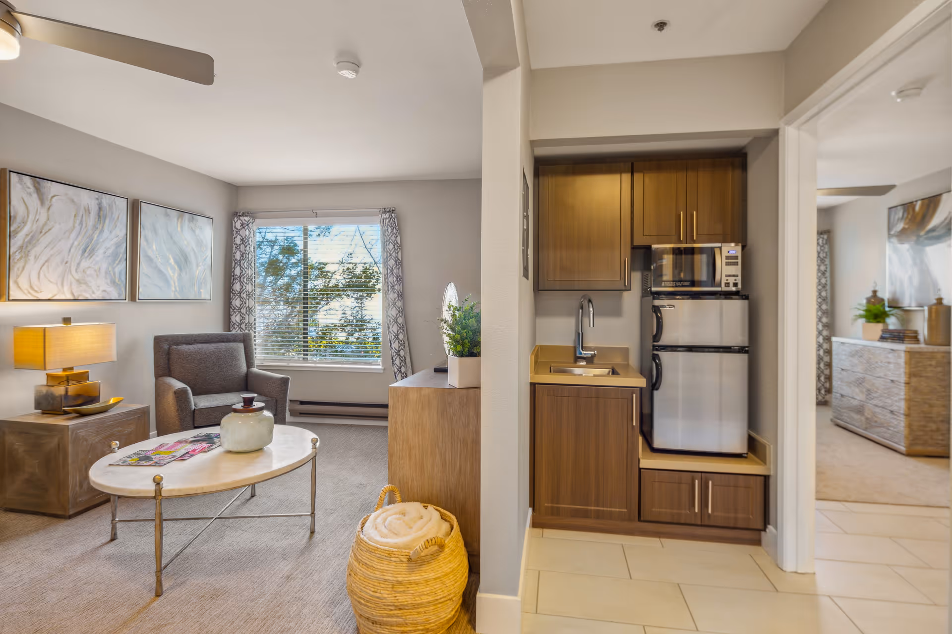 A cozy living area with a gray armchair, a round coffee table with a decorative jar and magazines, a side table with a lamp, and two abstract paintings on the wall. Adjacent to the living area is a small kitchenette with wooden cabinets, a sink, a microwave, and a mini refrigerator. A window with patterned curtains lets in natural light, and a woven basket with a rolled blanket is on the floor.