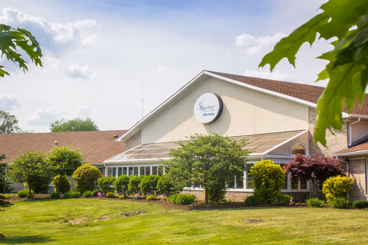 Front exterior of a single-story senior living facility with a glass sunroom, landscaped shrubs and a round 'Signature' sign on the facade.